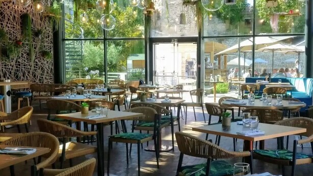 An outdoor dining area with wicker furniture, tables, and chairs, and large windows offering a view of the street. - Aquabella Hôtel & Spa (Aix-en-Provence)