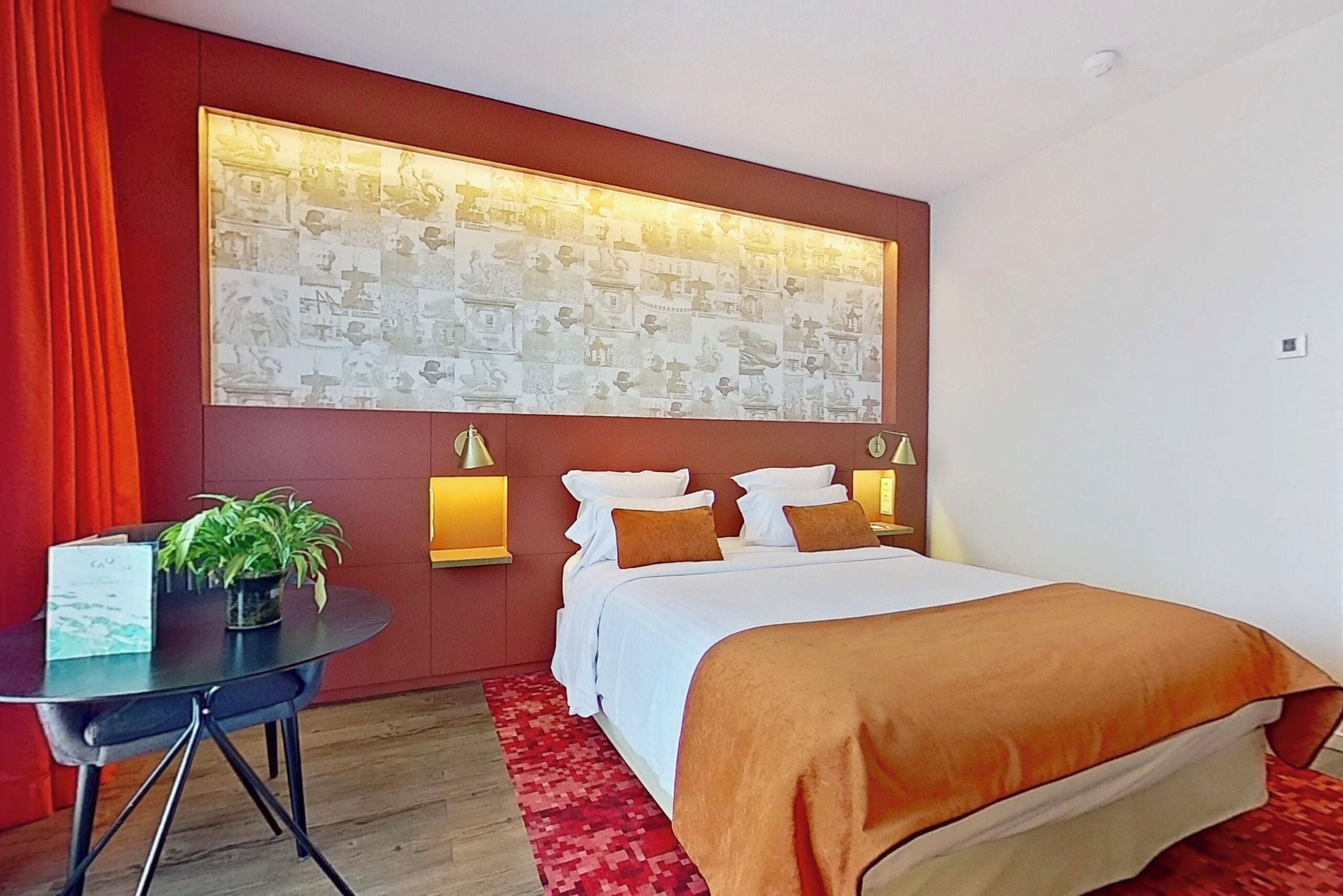 Een hotelkamer met een groot bed, een bureau met een plant, een rood gordijn en een muur met een kunstwerk met een patroon.