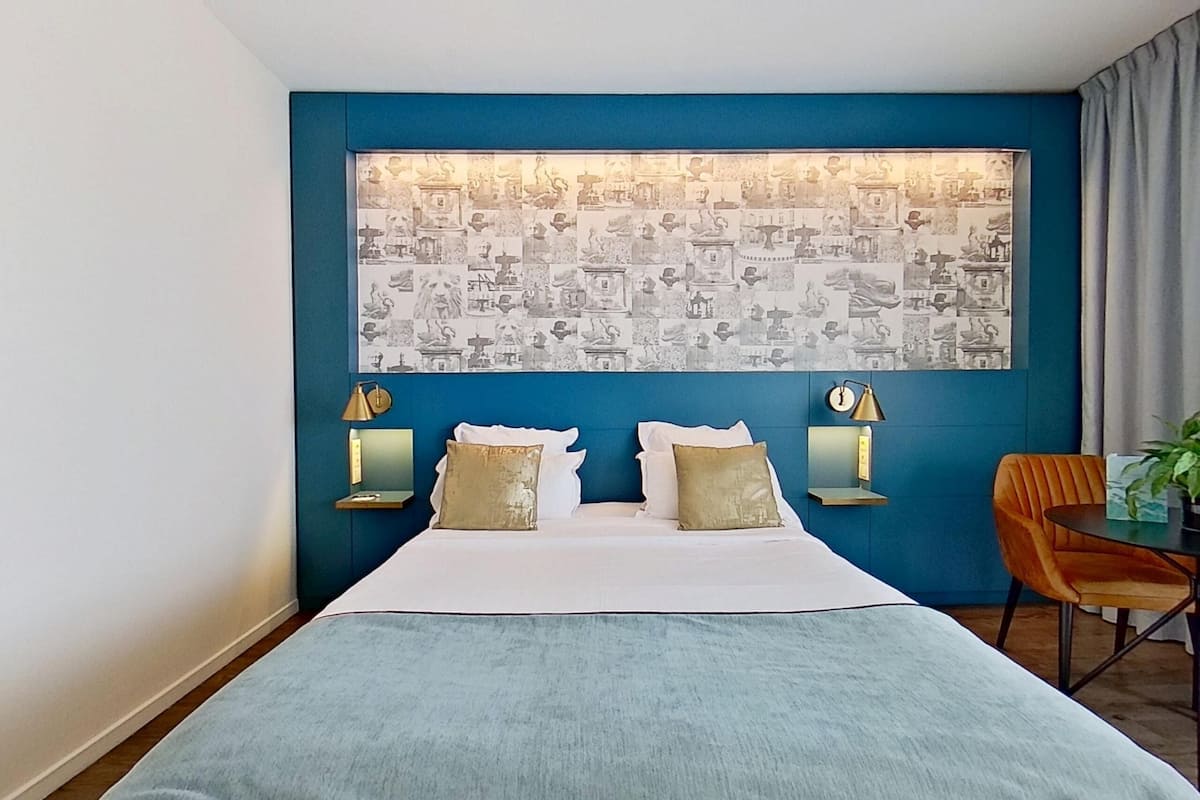 Une chambre d’hôtel avec un grand lit, deux tables de chevet avec des lampes, un mur d’accent bleu et une tapisserie à motifs.