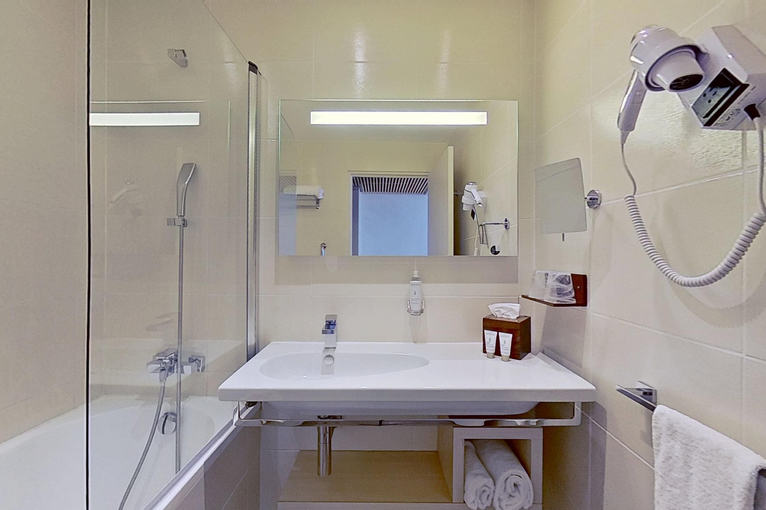 Une salle de bain moderne dotée d’une douche à parois de verre, d’un lavabo blanc, d’un miroir et d’un sèche-cheveux fixé au mur.