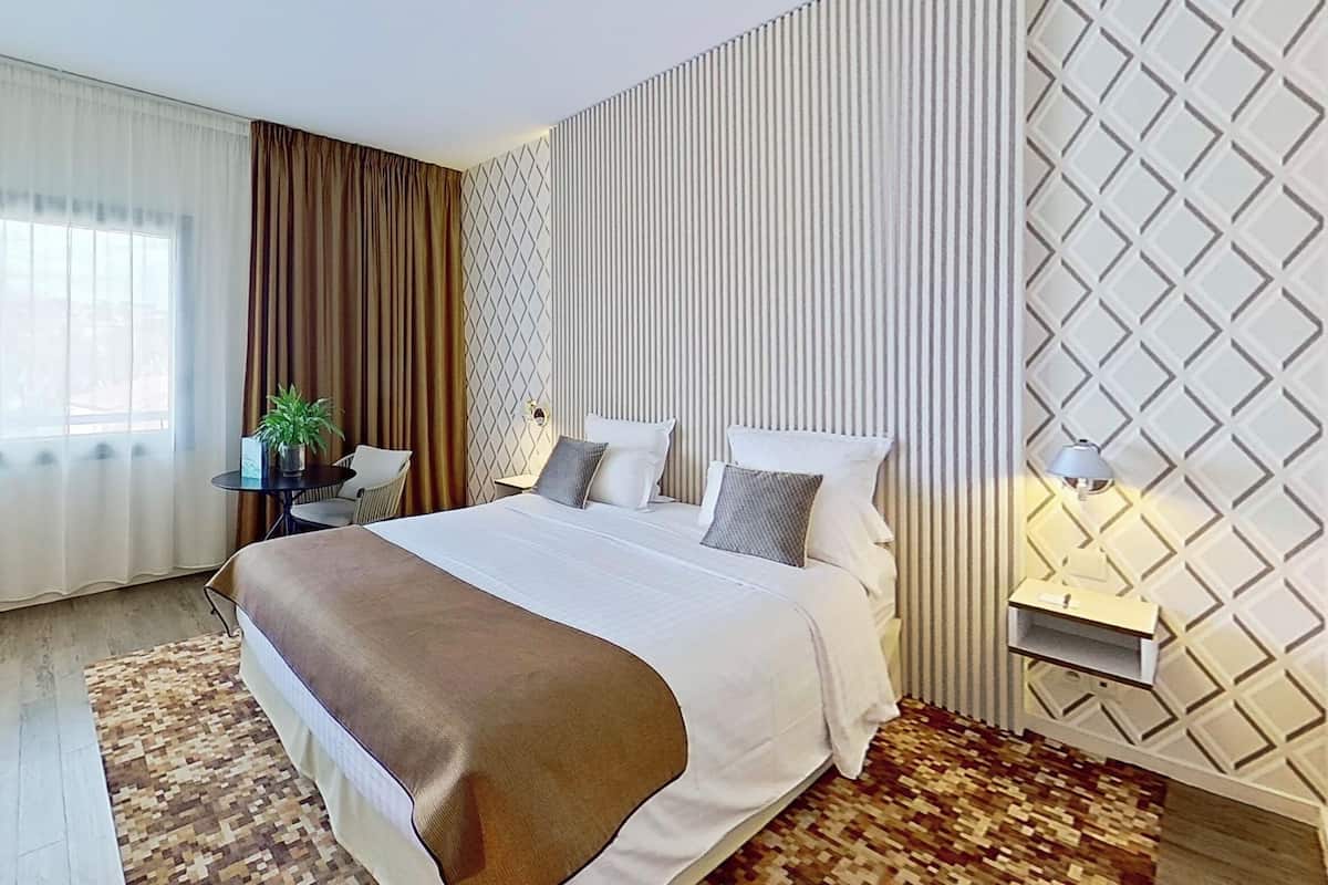 Une chambre d’hôtel avec un grand lit, une petite table sur laquelle est posée une plante, une lampe de chevet et un mur orné d’un motif.