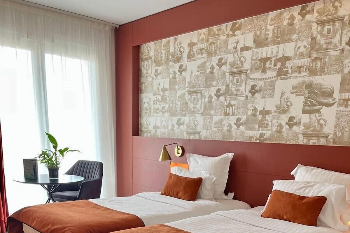 Une chambre d’hôtel avec deux lits, une moquette rouge, un mur orné d’une fresque à motifs, une petite table avec une plante et une chaise.