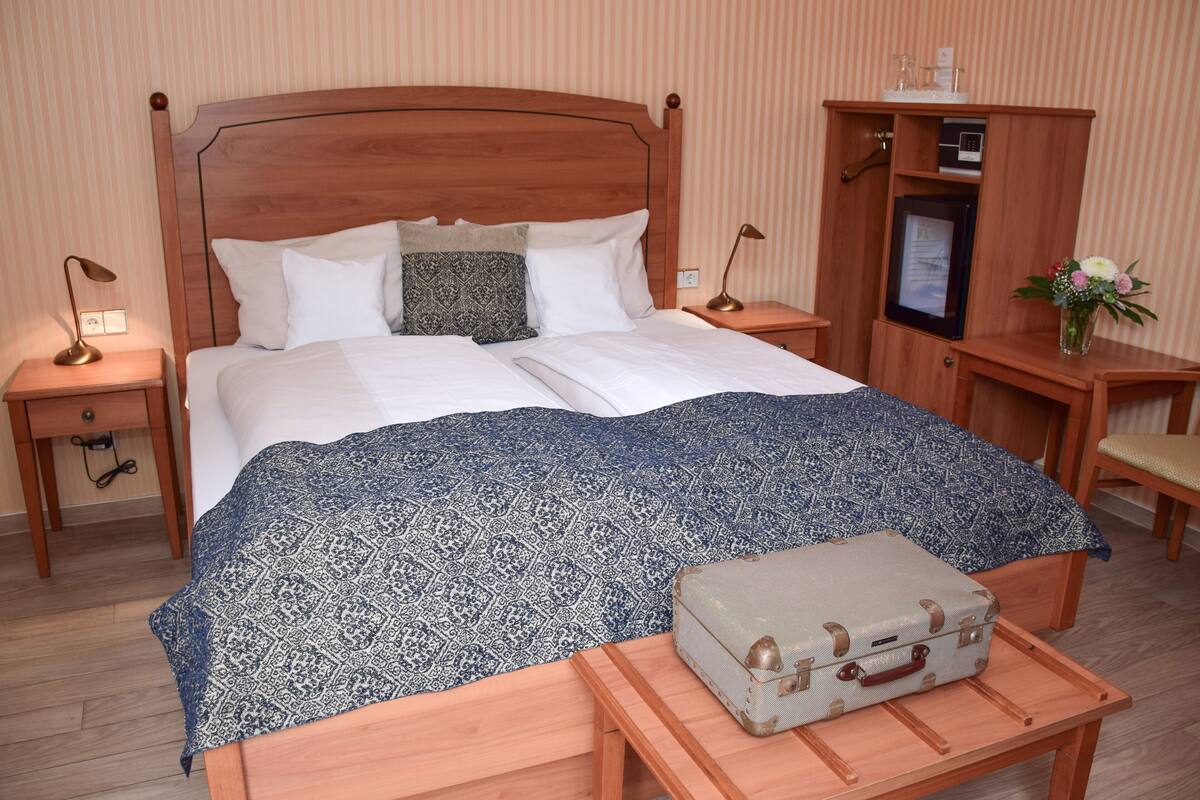 A neatly made bed with a patterned bedspread, a wooden headboard, and a bedside table with a lamp. A suitcase is placed on a small table beside the bed.