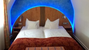 A room with a bed under a blue-lit arch, two bedside tables, and a floral-patterned bedspread. - Hotel Barbara (Freiburg im Breisgau)
