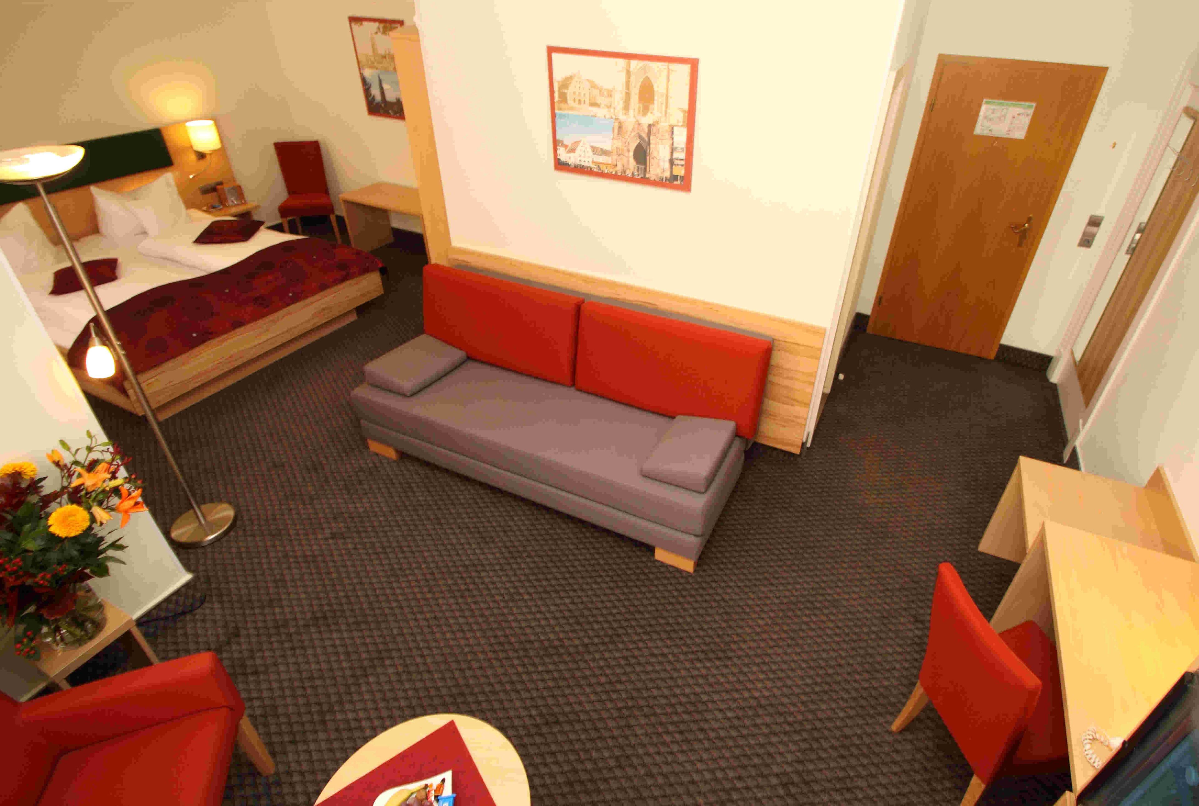 A hotel room with a bed, a sofa, a small table with a vase of flowers, a chair, and a door.