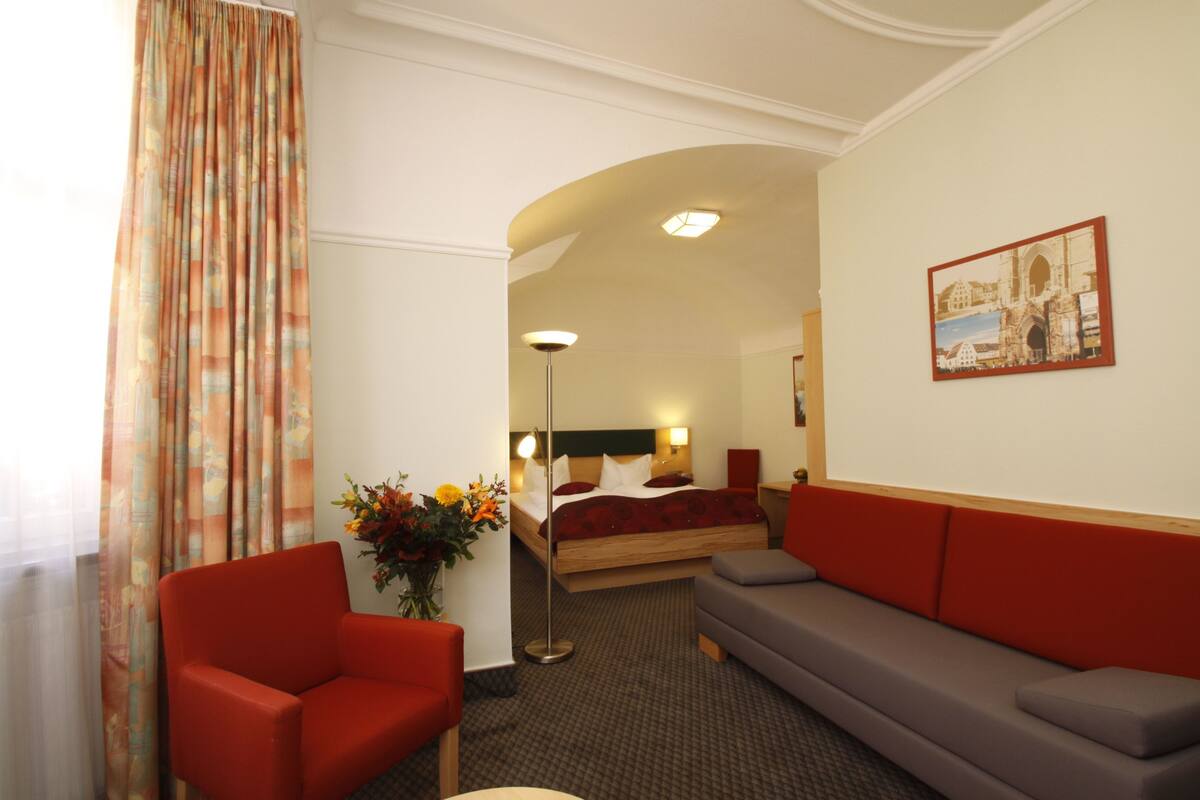 A hotel room with a bed, a red armchair, a grey sofa, a floor lamp, and a painting on the wall.