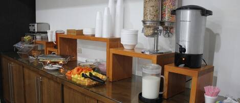 A buffet setup with various food containers, a milk dispenser, and stacked white cups.