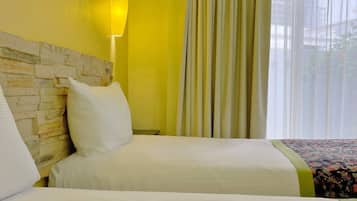 Une chambre d’hôtel avec deux lits, une tête de lit à l’aspect pierre, une lampe fixée au mur et une fenêtre ornée de rideaux légers.