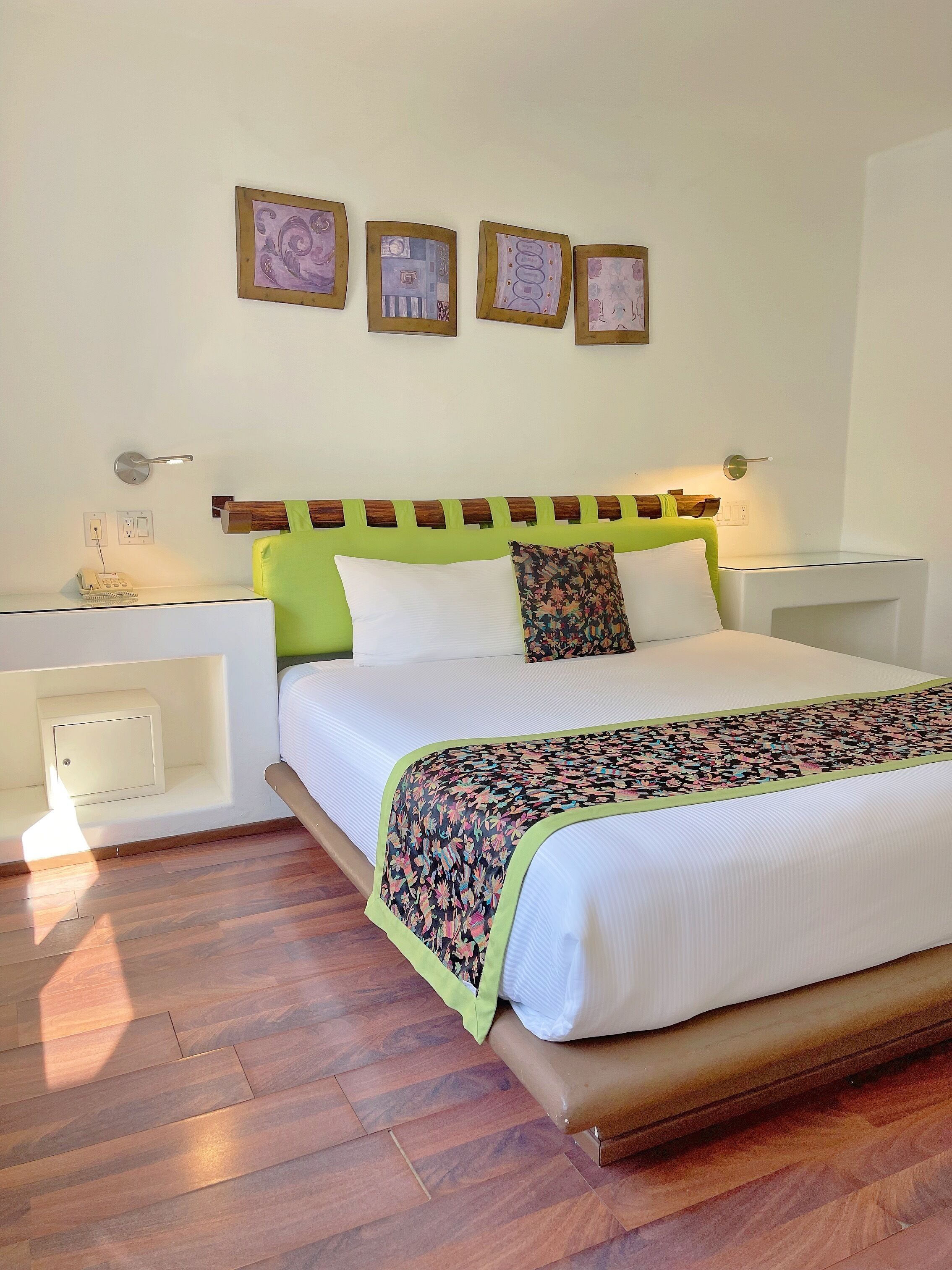 A hotel room with a bed, bedside tables, a nightstand, a wall-mounted telephone, and three framed artworks.