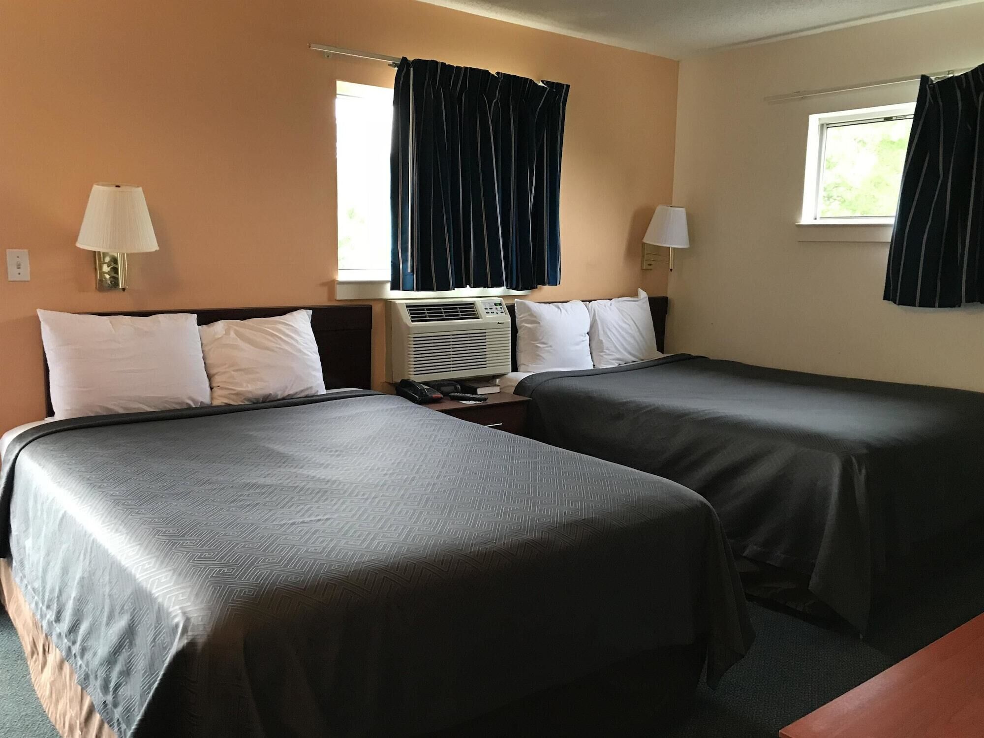 A hotel room with two beds, a window with curtains, an air conditioning unit, and a nightstand with a lamp.