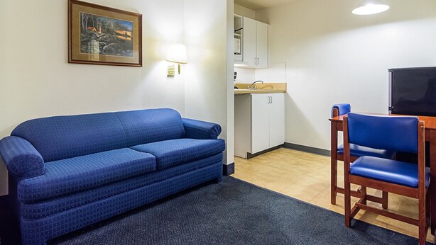 A compact room with a blue sofa, a wooden chair, a small table, a painting on the wall, and a kitchenette in the background. - Motel 6 - Fayetteville, NC - Fort Liberty Area (Fayetteville)