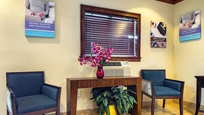 A reception area with two blue chairs, a wooden console table with a vase of pink flowers, and a large window with blinds. - Motel 6 - Fayetteville, NC - Fort Liberty Area (Fayetteville)