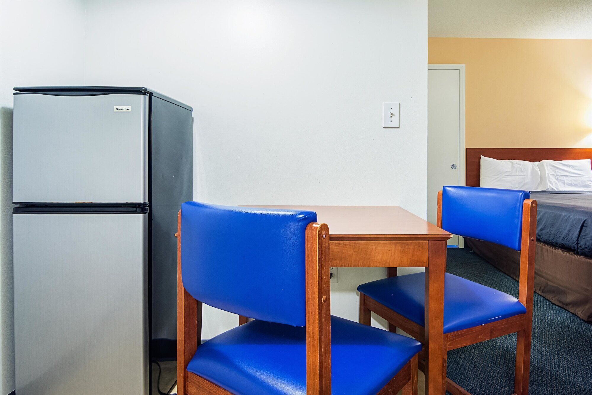 A compact hotel room with a kitchenette, a wooden table, two blue chairs, and a bed with a headboard.