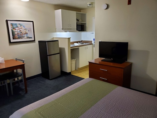 Habitación de hotel pequeña y funcional con cama, escritorio, refrigerador, televisión, microondas y kitchenette.