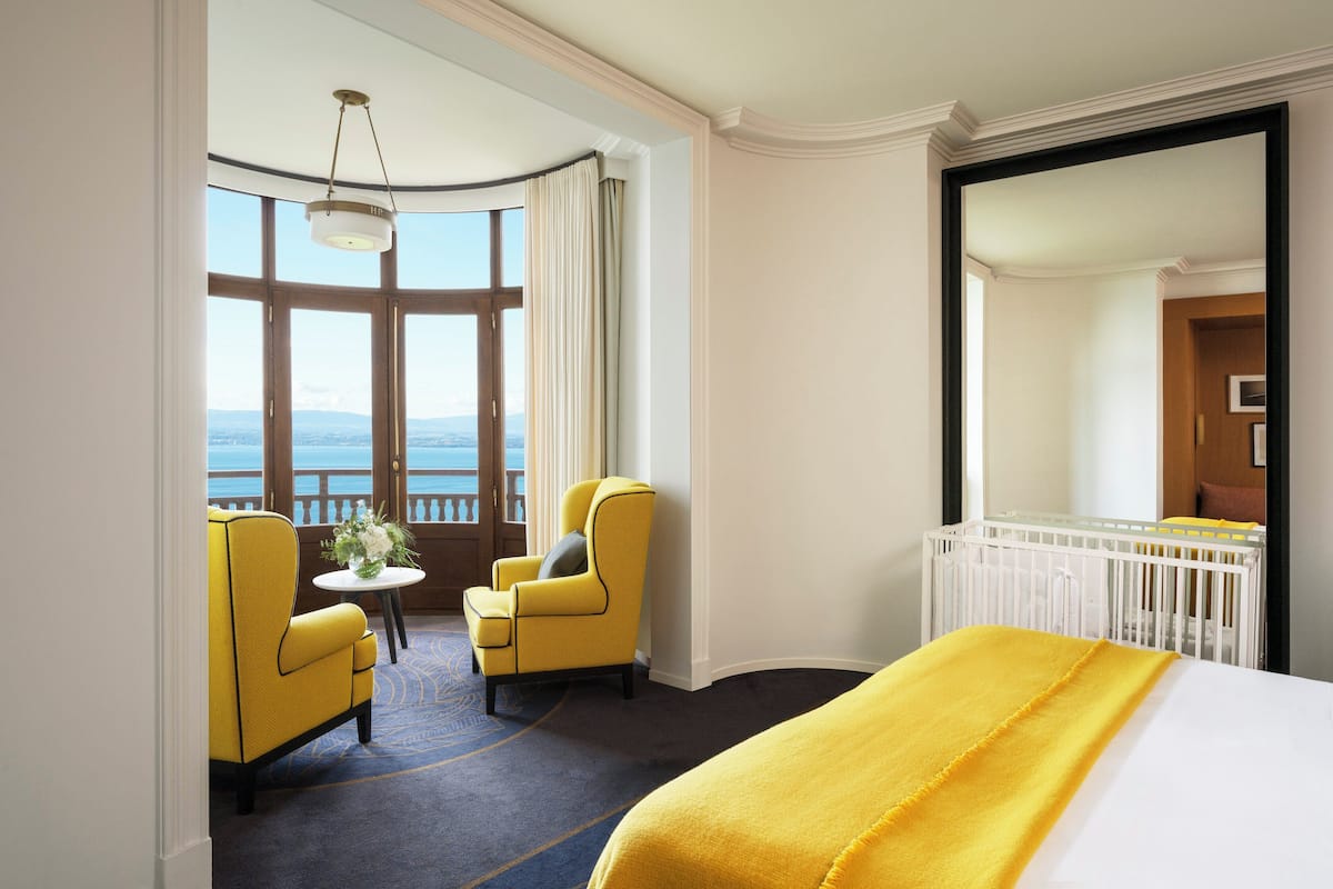 Een hotelkamer met een groot bed, uitzicht op zee, twee gele fauteuils, een klein tafeltje met een vaas en een wit ledikant.