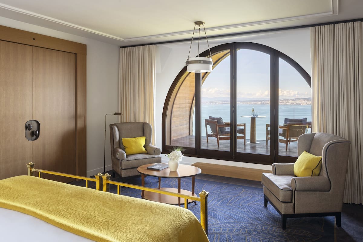 Een hotelkamer met een groot raam, een bed, twee fauteuils, een kleine tafel en uitzicht op zee.