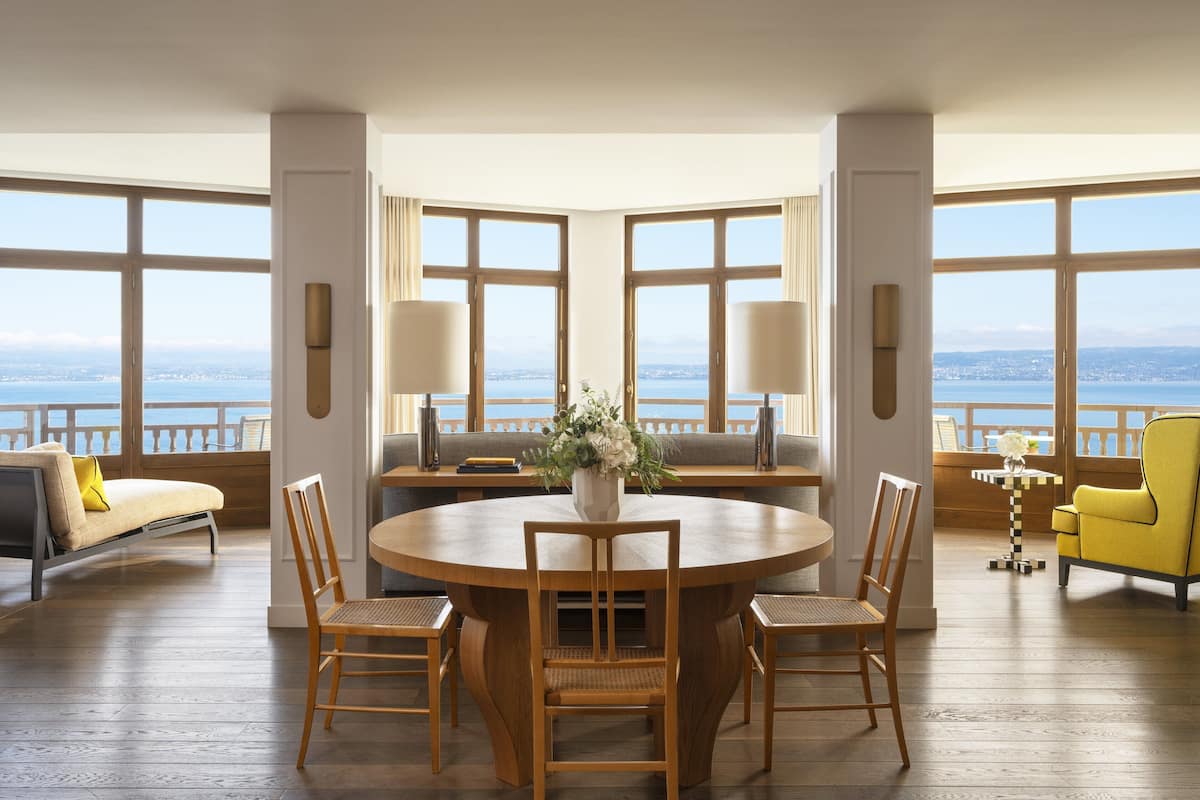 Ruime kamer met een ronde houten tafel, vier stoelen, een bank en een gele fauteuil. Grote ramen bieden uitzicht op zee.