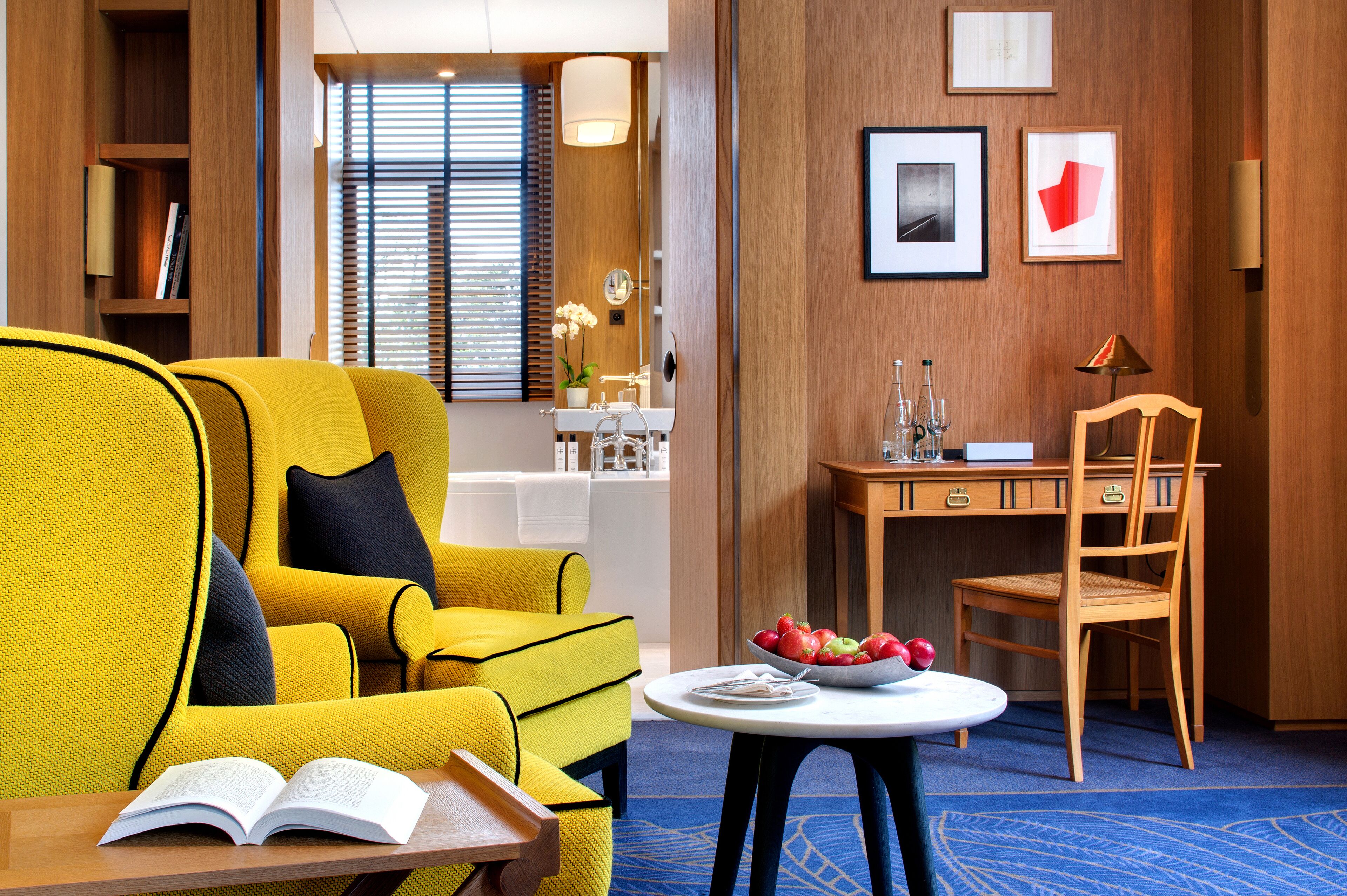 A hotel room with a yellow armchair, a small round table with a bowl of fruit, a wooden desk, and a chair.