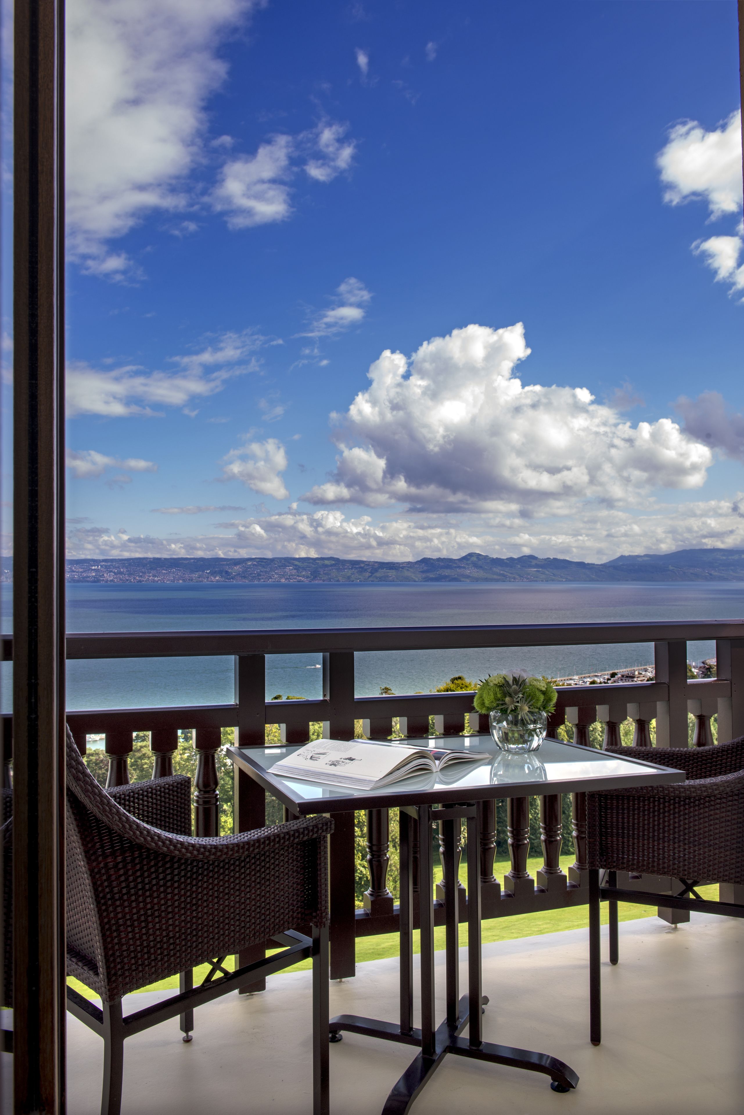 Un balcón con sillas de mimbre y una mesa, con vista a un paisaje marino y montañoso.