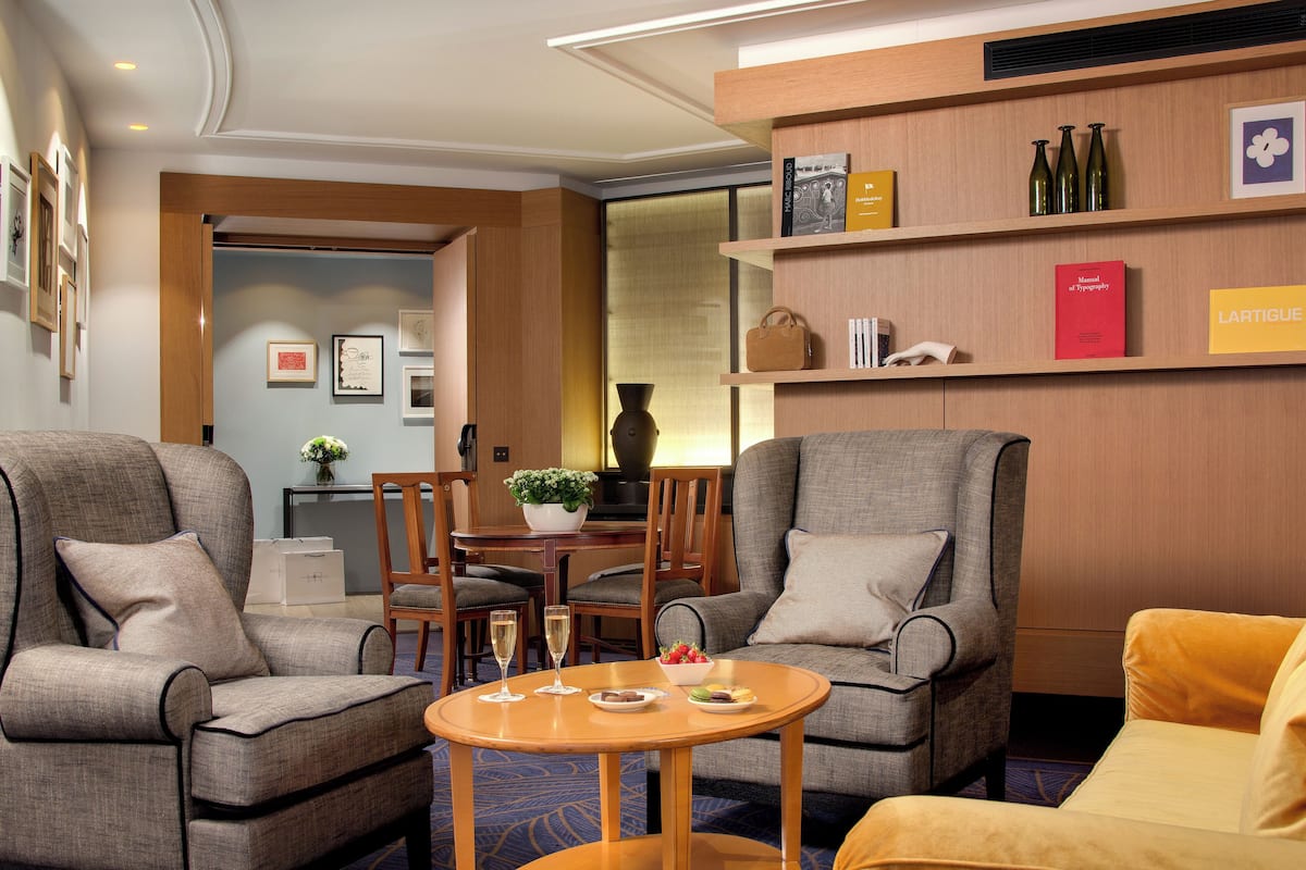 Een hotelkamer met twee fauteuils, een bank, een eettafel met een vaas en fruit, en een boekenkast met boeken en decoratieve objecten.