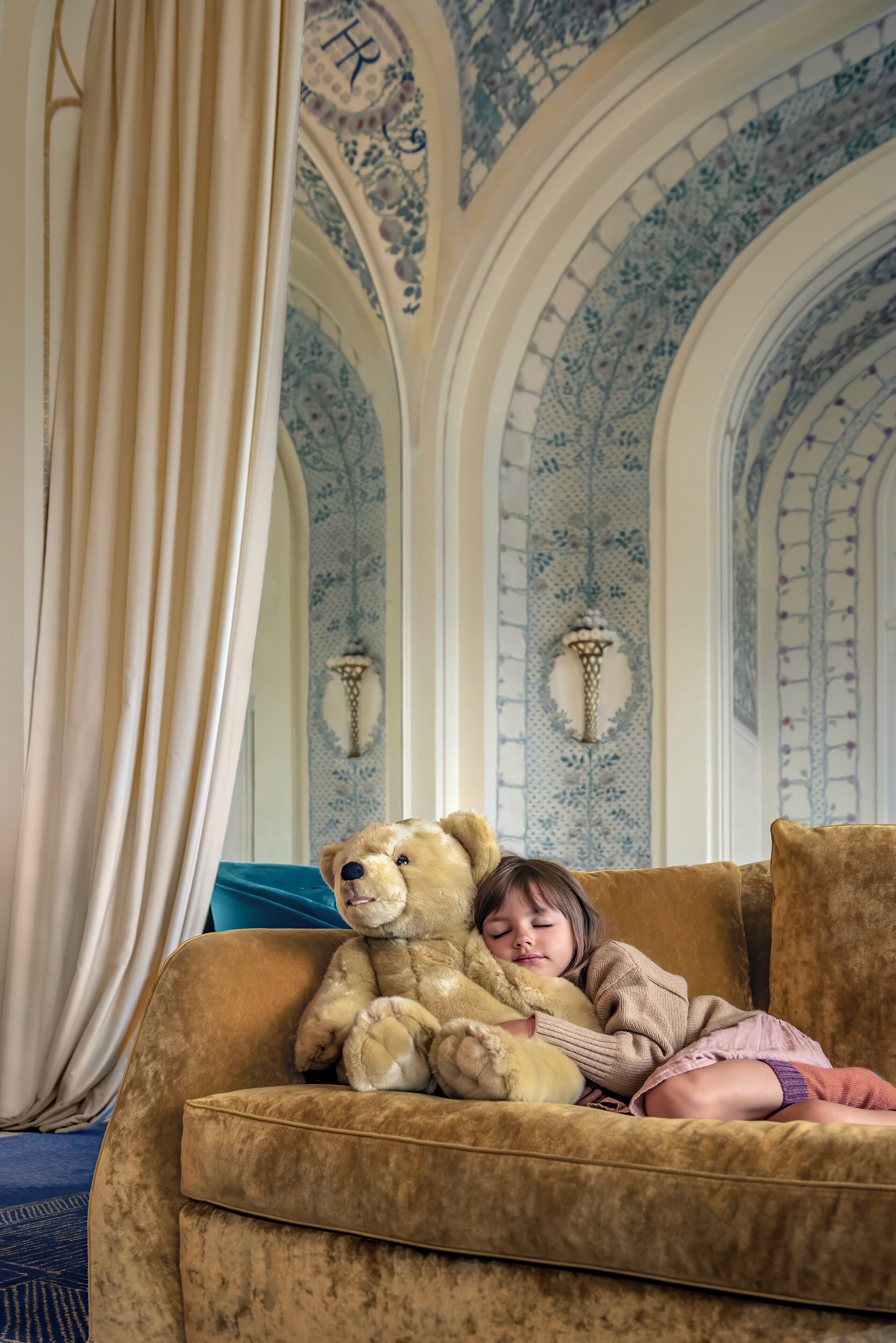Un niño duerme en un sofá con un gran oso de peluche a su lado, en una habitación con paredes de intrincado diseño azul y blanco y un candelabro.
