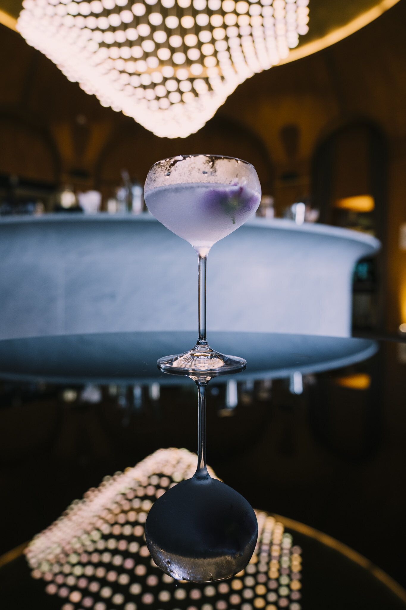 A martini glass with a purple drink on a bar counter with a decorative light fixture above.