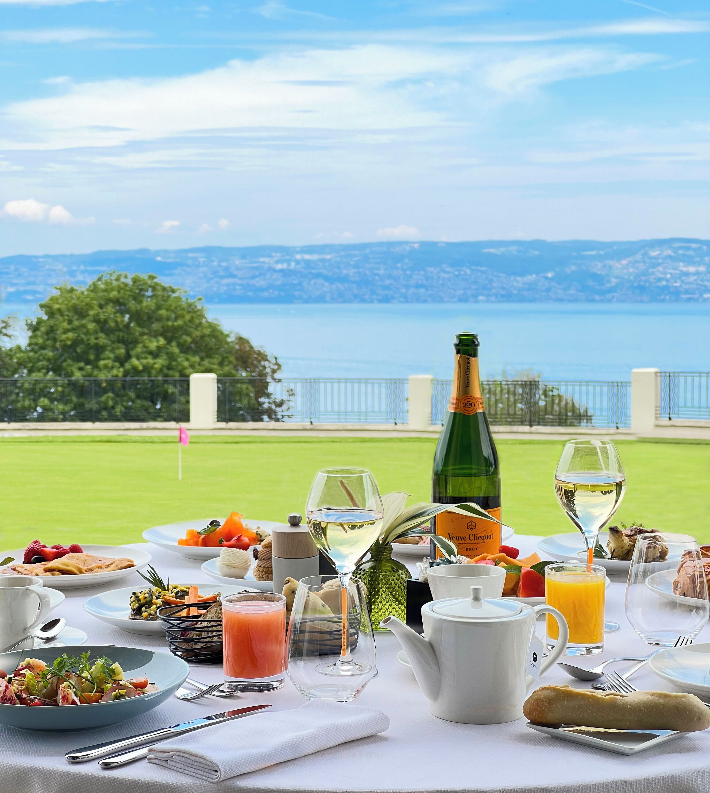 Ein Tisch, reichhaltig gedeckt mit Frühstücksleckereien wie Obst, Gebäck und Getränken, mit Blick auf einen malerischen See und Stadtpanorama.