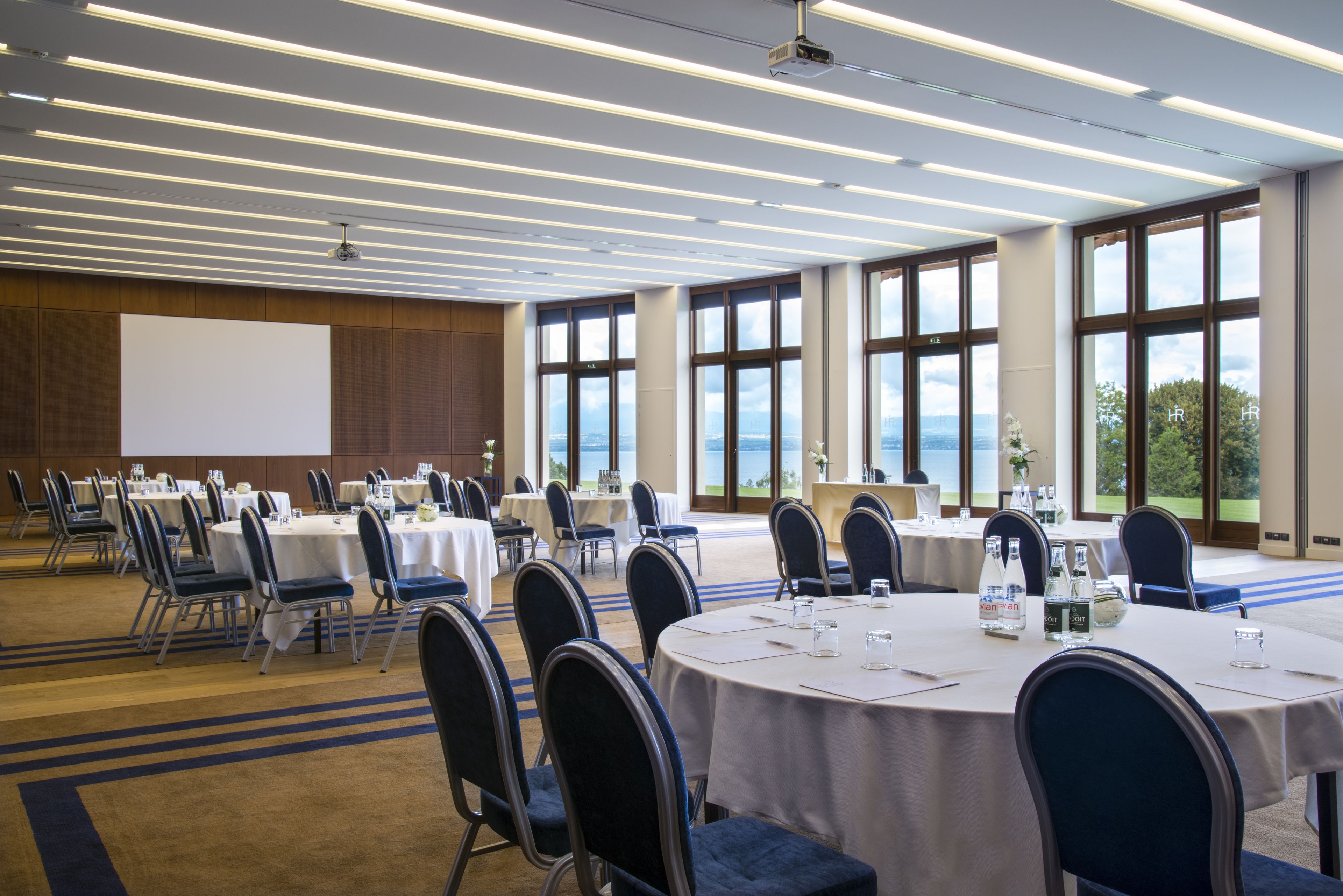 Ein Konferenzraum mit runden Tischen, Stühlen und einem großen Bildschirm.