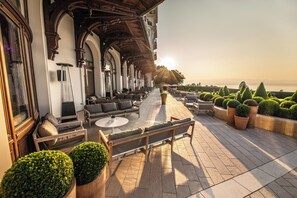 A terrace with outdoor seating, a view of the sea, and a building with arched windows and a wooden ceiling. - Hôtel Royal (Evian-les-Bains)
