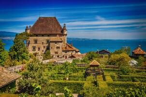A historic stone building with multiple towers, surrounded by lush greenery and vineyards, overlooking a body of water.