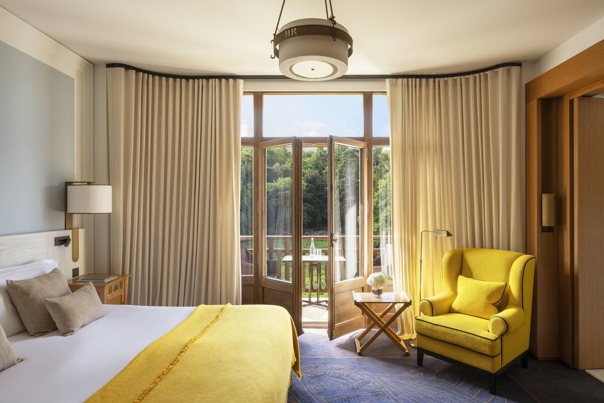 Hotelkamer met een groot bed, een gele fauteuil, een balkon met een tafel en stoelen, en uitzicht op groen.