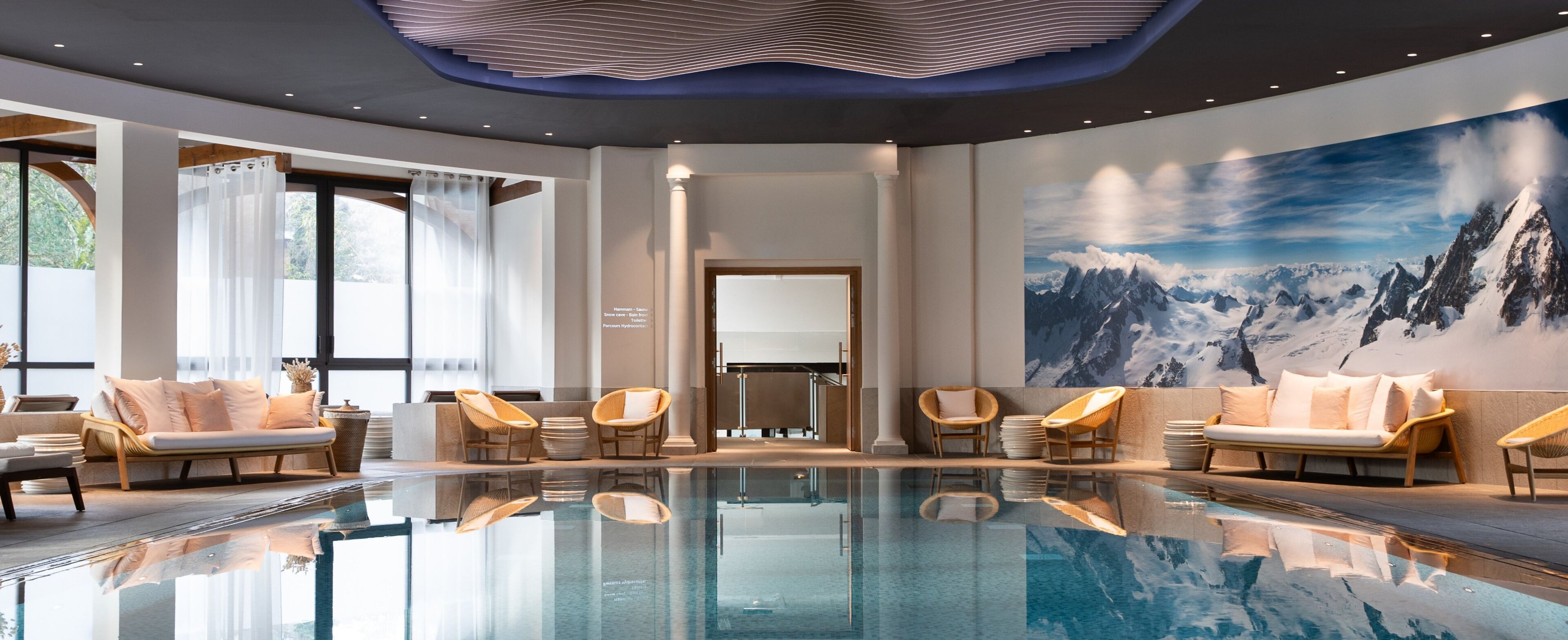 A modern indoor pool area with clear glass walls, comfortable seating, and a large mountain landscape mural on the wall.