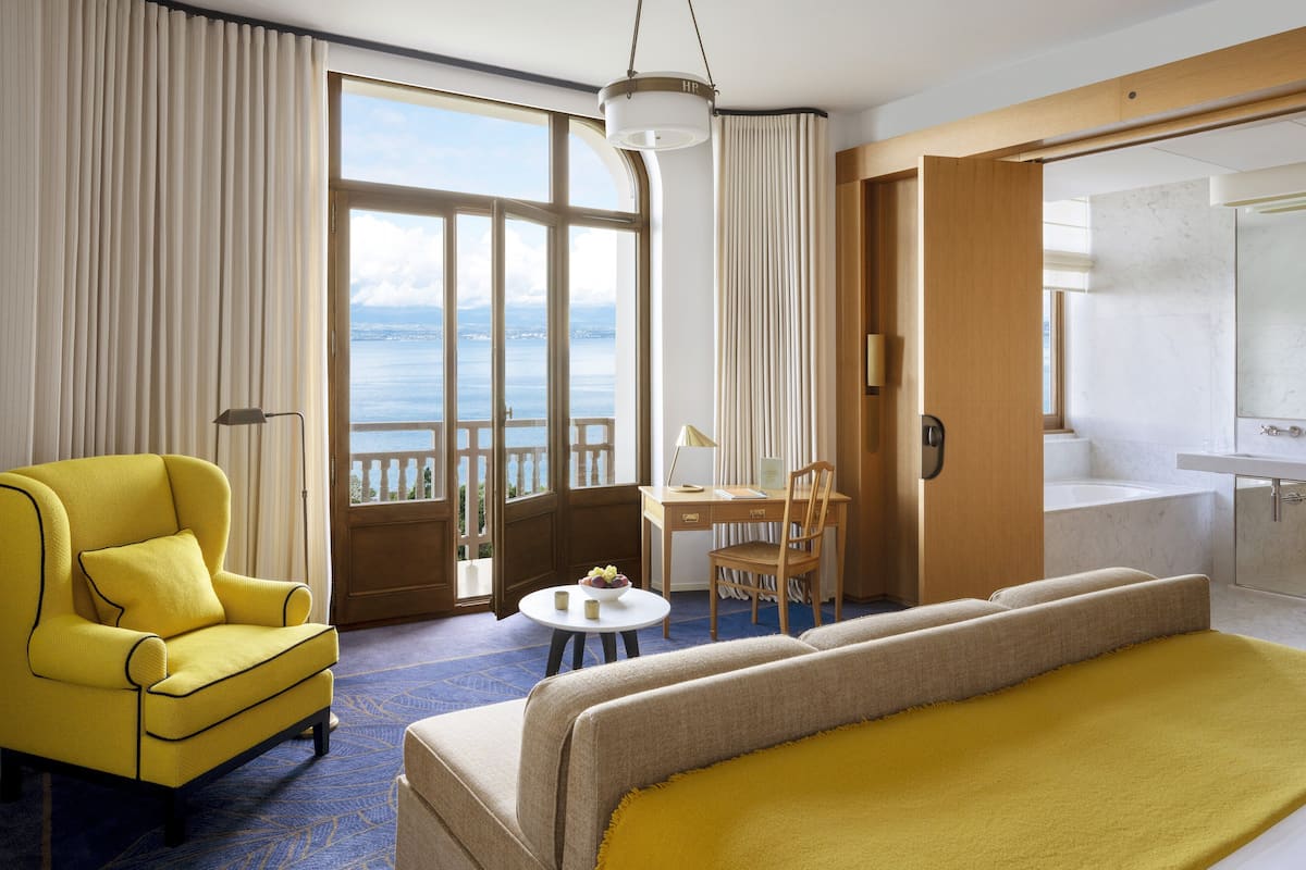 Hotelkamer met een bed, een gele fauteuil, een bureau met stoel, een tafeltje met een schaal fruit, en een badkamer met bad en wastafel.