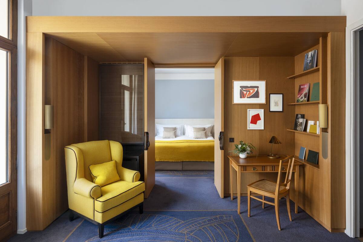 Een kleine hotelkamer met een gele fauteuil, een bed met een geel dekbed, een bureau met stoel, en een boekenplank met ingelijste foto’s.