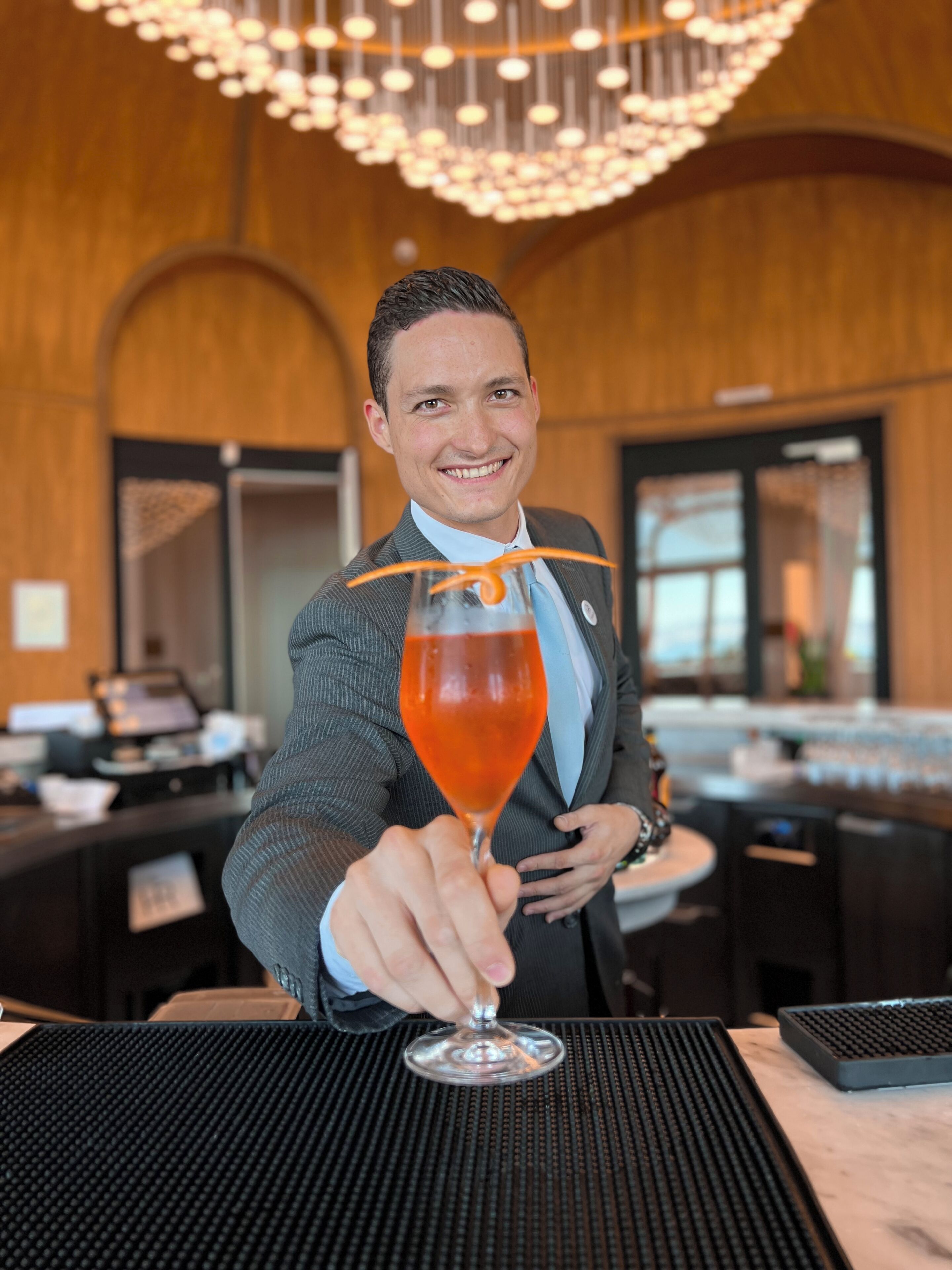 Una persona con traje sostiene una copa de cóctel adornada con una rodaja de naranja. El lugar parece un bar o restaurante con decoración de madera y una lámpara de araña moderna.
