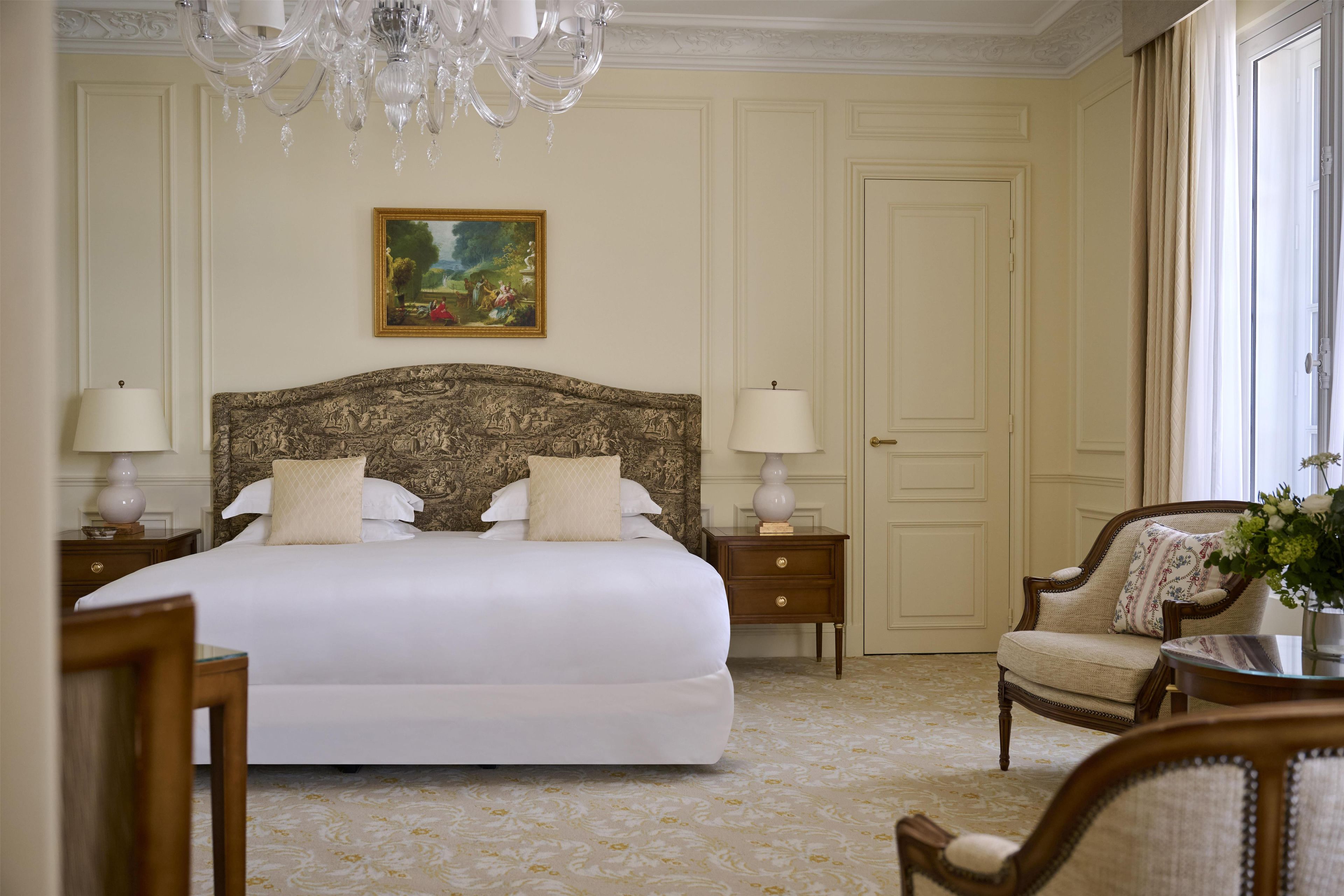 A hotel room with a large bed, two bedside tables, a chair, a chandelier, and a door.