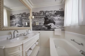 A bathroom with a large mirror, two sinks, a bathtub, and a mural of a coastal town.