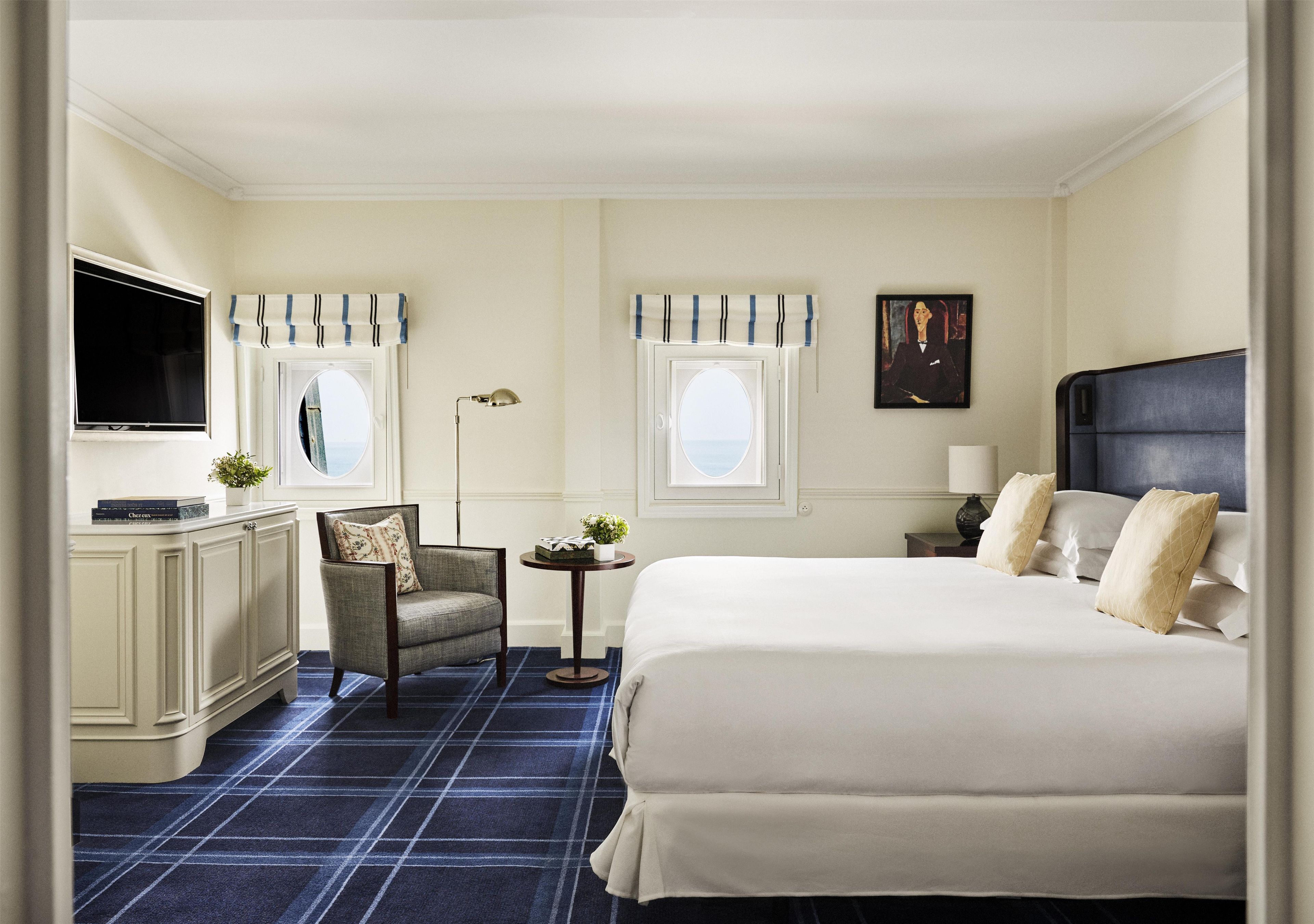 A hotel room with a bed, a chair, a desk, a television, and two windows with curtains.