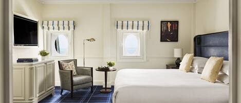 A hotel room with a bed, a chair, a desk, a television, and two windows with curtains.
