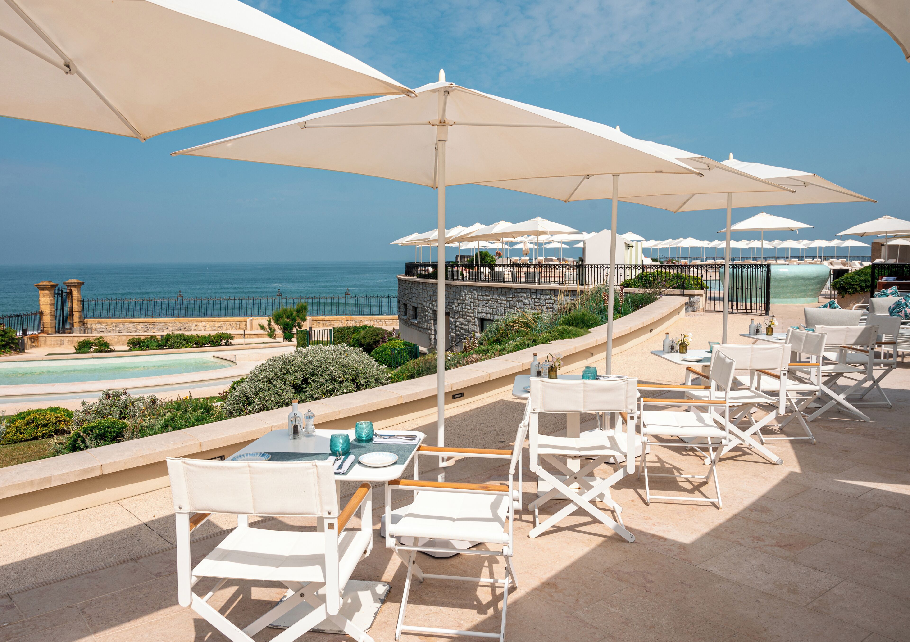 Terraza con mobiliario exterior blanco y sombrillas, con vistas a una piscina y al océano.