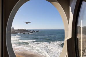 A view through a window of a coastal scene with waves, a sandy beach, and rocky outcrops.