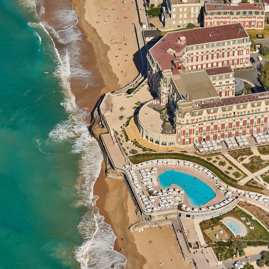 An aerial view of a coastal hotel with a beachfront, a swimming pool, and a parking area.
