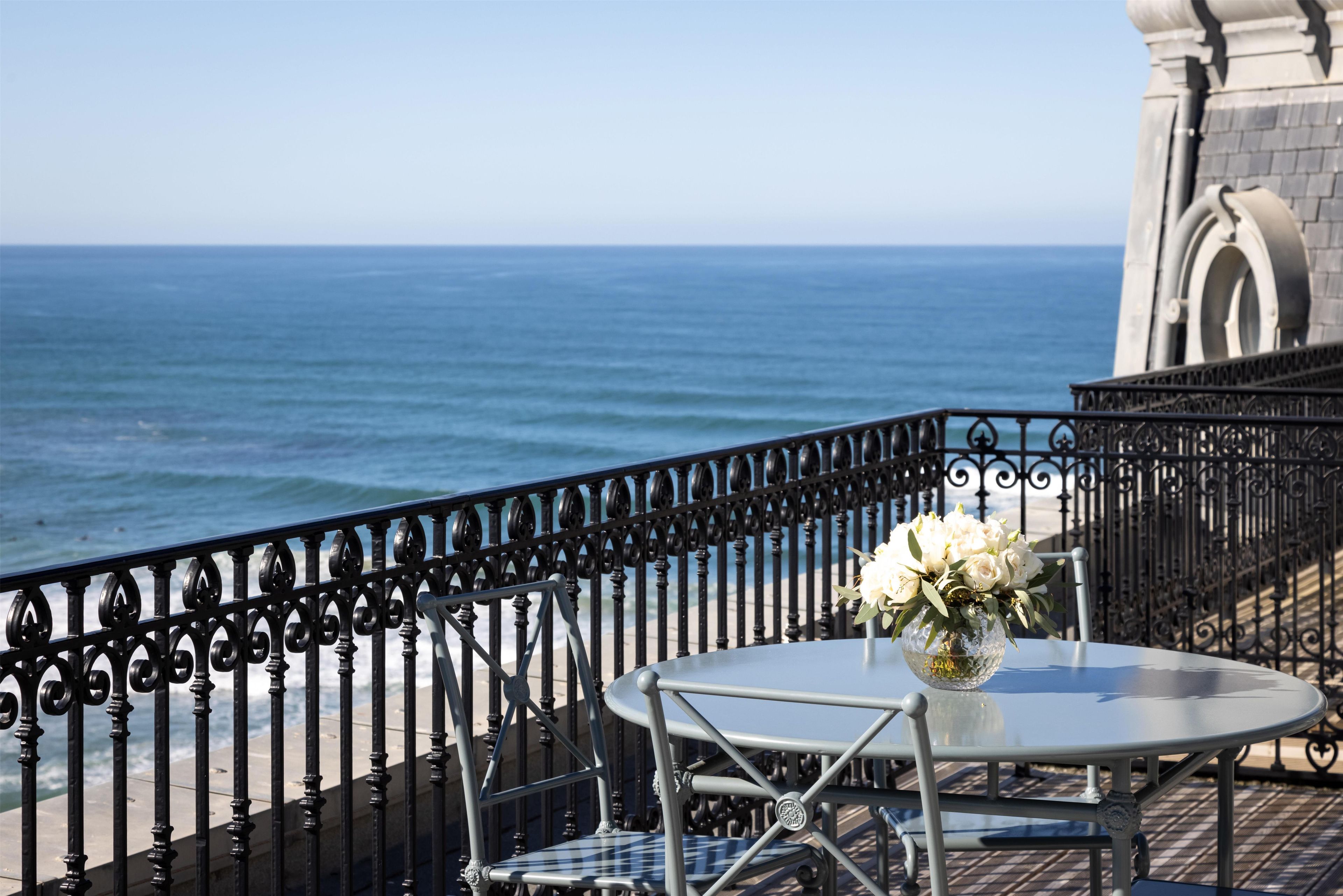A balcony with a metal railing, a table with a vase of flowers, and a view of the ocean.