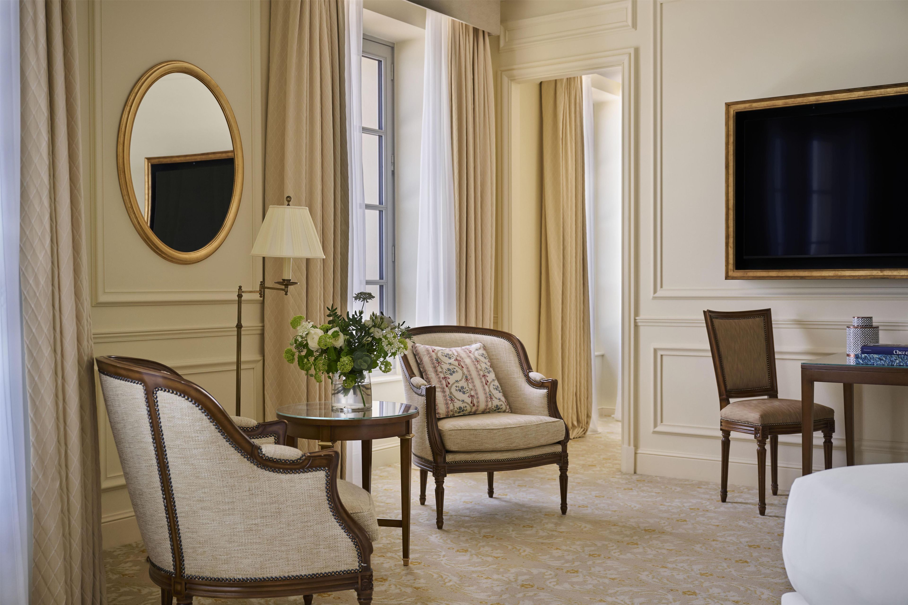 A hotel room with a large mirror, a table with a lamp, two armchairs, a sofa, and a television.