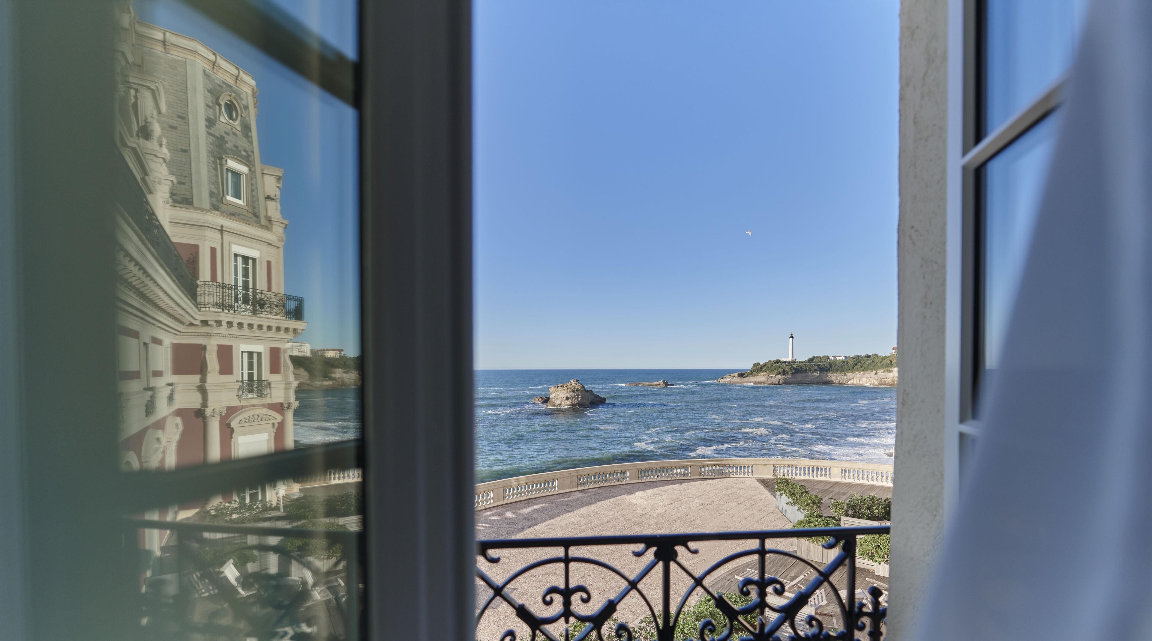 A balcony with a view of the sea, a building with classical architecture, and a lighthouse in the distance.