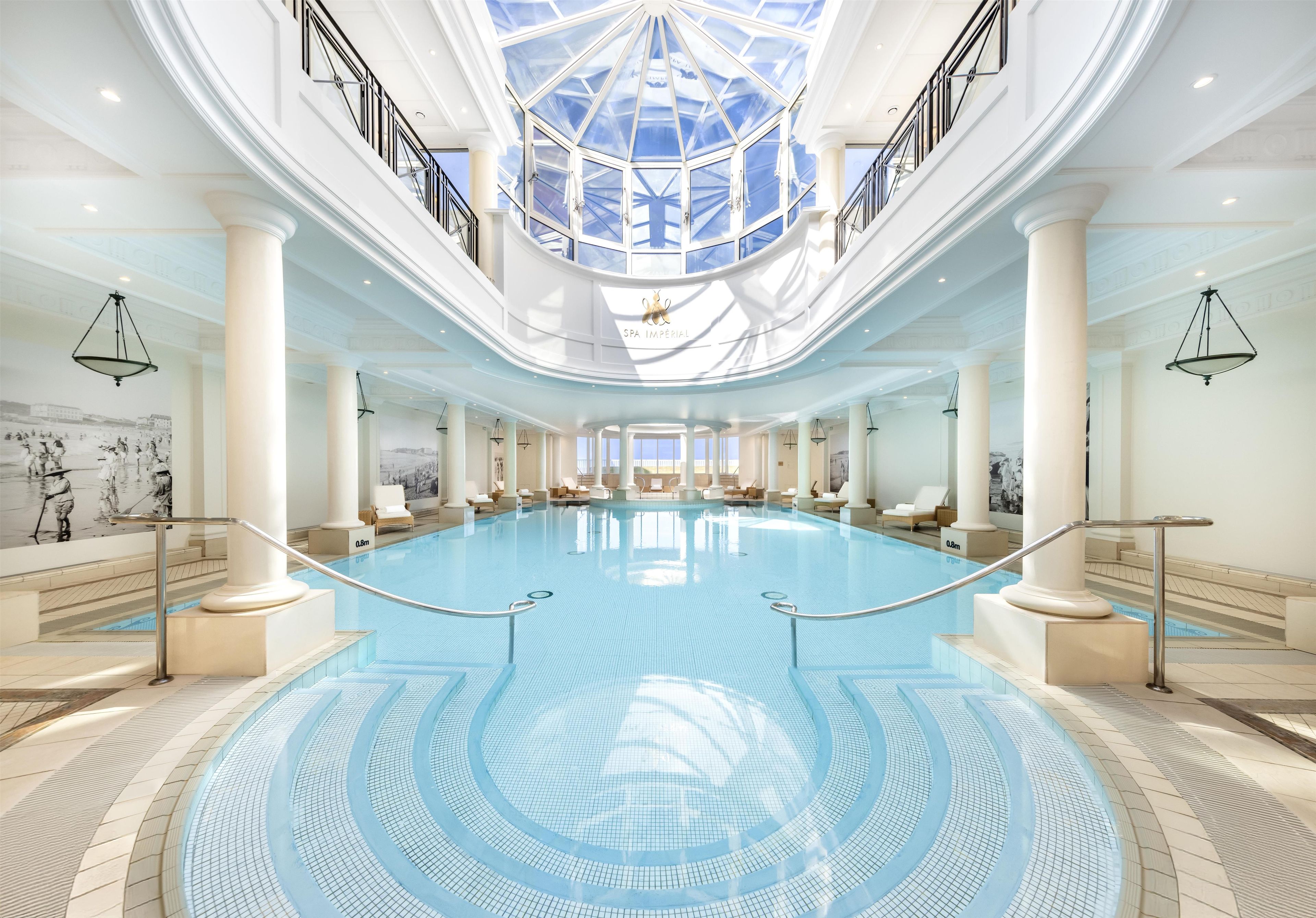 Una amplia piscina cubierta con techo de vidrio, rodeada de columnas y sillones de descanso.