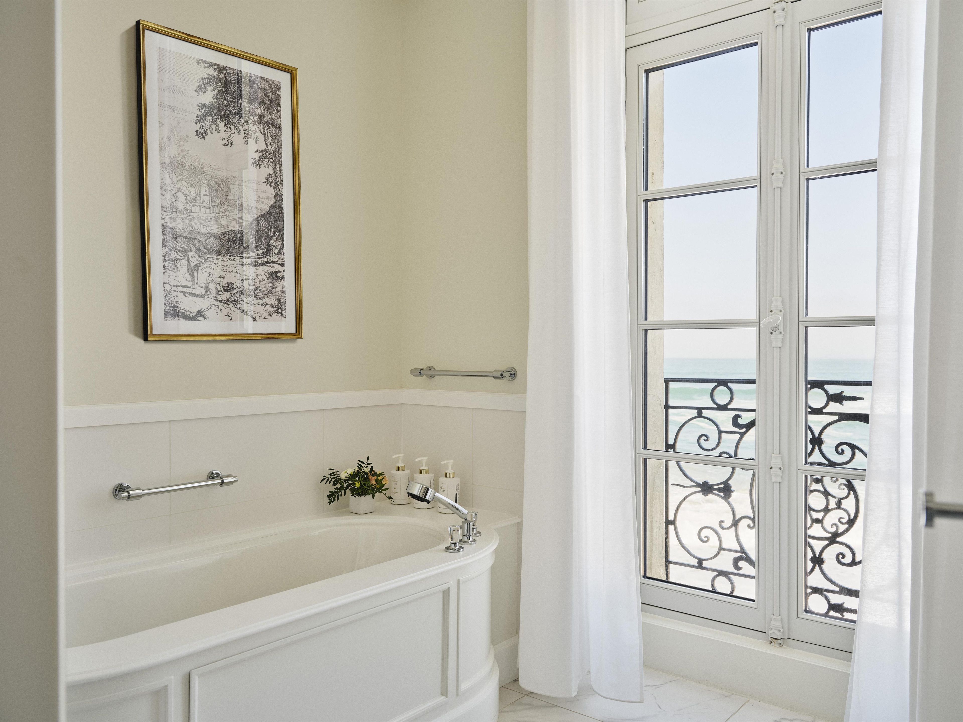A bathroom with a bathtub, a framed picture on the wall, and a window with a balcony view.