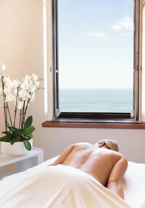 A room with a bed, a window with a view of the sea, and a potted orchid plant.