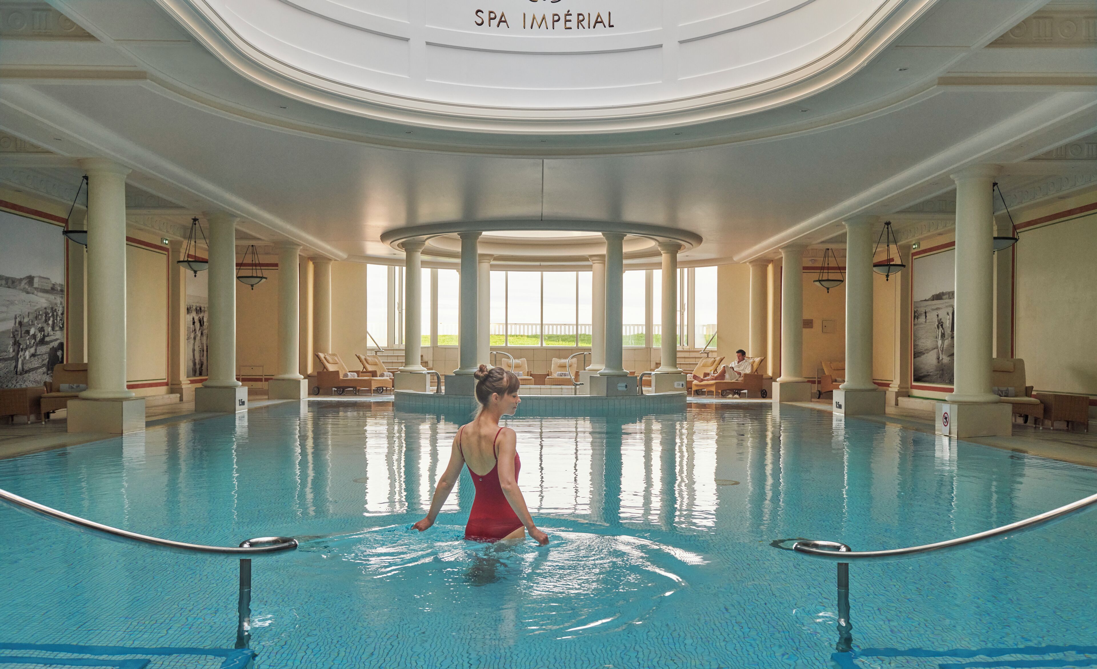 A large indoor swimming pool with a central circular area, lounge chairs, and a view of the outdoors.