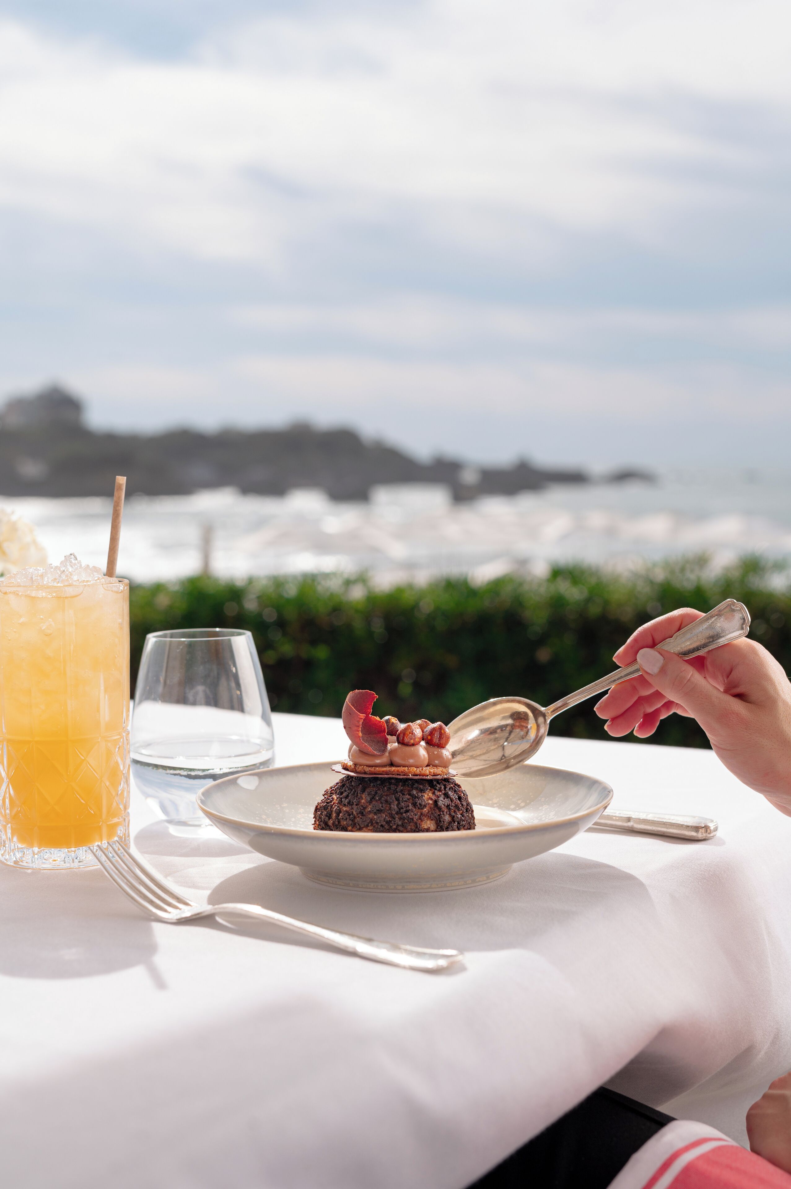 A dessert on a plate with a spoon, a glass of orange drink, and a glass of water on a table with a view of the ocean.